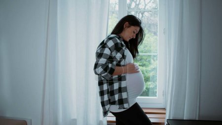 Glückseliger mütterlicher Moment, werdende Mutter streichelt 8-Monats-Bauch und erwartet Neugeborenes zu Hause am Fenster