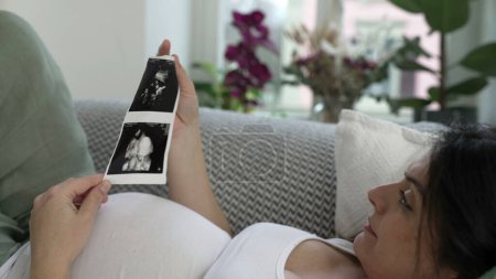 Foto de Mujer embarazada mira la imagen de ultrasonido de su bebé durante la etapa final del embarazo acostado en el sofá descansando en casa abrazando la alegría materna - Imagen libre de derechos