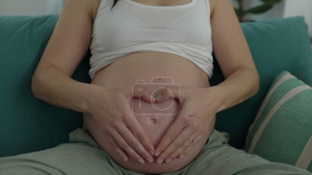 Téléchargez les photos : Maternité sincère - Joyeuse femme enceinte dans la trentaine assise sur le canapé à la maison, ce qui rend le symbole du cœur avec les mains sur le ventre pendant la grossesse tardive - en image libre de droit