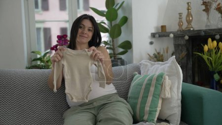 Maternal Anticipation - Expectant Mother in Late Pregnancy Showing Baby Clothes to camera, Comfortably Seated on Couch, Embracing the Joys of Upcoming Motherhood