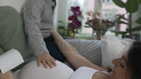 Foto de Madre e hijo compartiendo un momento feliz y tierno mientras miran el ultrasonido del hermano bebé mientras descansan en el sofá durante el embarazo en etapa tardía. Niño tocando suavemente el vientre - Imagen libre de derechos