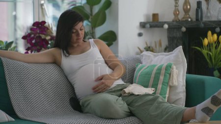 Foto de Mujer embarazada acariciando el vientre sentada en el sofá en casa durante el embarazo del tercer trimestre. Señora de los años 30 anticipando al bebé recién nacido durante la etapa final - Imagen libre de derechos