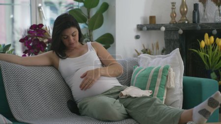 Foto de Mujer embarazada acariciando el vientre sentada en el sofá en casa durante el embarazo del tercer trimestre. Señora de los años 30 anticipando al bebé recién nacido durante la etapa final - Imagen libre de derechos
