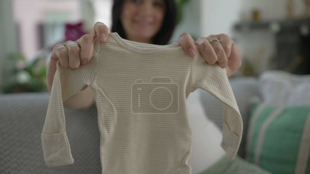 Téléchargez les photos : Anticipation maternelle - La future mère en fin de grossesse affiche fièrement les vêtements de bébé à la caméra, confortablement assis sur le canapé, rayonnant de joie et d'excitation pour la maternité à venir - en image libre de droit