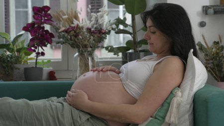 Foto de Mujer embarazada descansando en el sofá acariciando el vientre en anticipación de una nueva vida creciendo dentro. Perfil de lady durign 30s tercer trimestre del embarazo - Imagen libre de derechos
