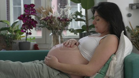 Foto de Mujer embarazada descansando en el sofá acariciando el vientre en anticipación de una nueva vida creciendo dentro. Perfil de lady durign 30s tercer trimestre del embarazo - Imagen libre de derechos
