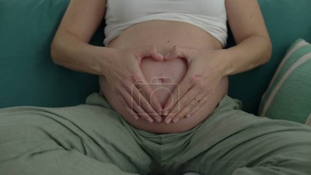Heartfelt Maternity - Joyful Pregnant Woman in Her 30s Seated on Couch at Home, Making Heart Symbol with Hands on Belly During Late Stage Pregnancy