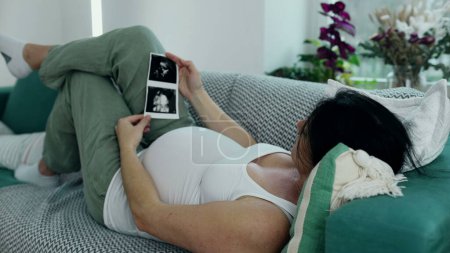 Photo for Pregnant woman laid on couch looking at ultrasound image of her baby during third trimester pregnancy. Expecting parent awaits her newborn - Royalty Free Image