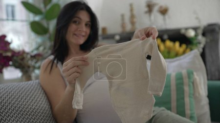 Téléchargez les photos : Moments de maternité joyeuse - Mère en fin de grossesse mettant en vedette de minuscules tenues de bébé à la caméra, détendu et souriant sur le canapé, incarnant le bonheur d'attendre un enfant - en image libre de droit