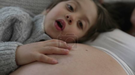 Mütterliche Liebe eingefangen - Sohn stützt sich als Zurschaustellung von Zuneigung und Fürsorge auf den Schwangerschaftsbauch der Mutter, Mutter ruht zu Hause auf Couch und wartet auf Neugeborenes