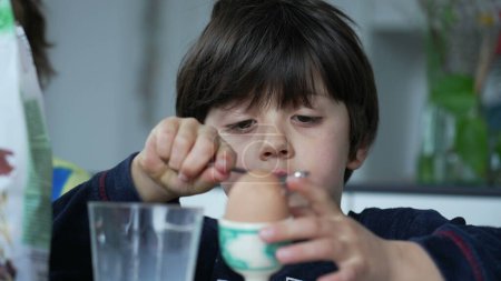 Niño rompiendo huevo abierto con cuchara en la mesa del desayuno. Un niño pequeño concentrado abriendo huevo hervido con cuchara