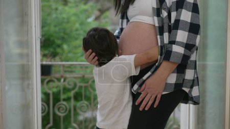 Niño cariñoso besándose y abrazando el vientre de la madre en el tercer trimestre del embarazo mostrando afecto hacia el hermano bebé nonato
