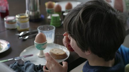 Foto de Parte posterior del niño comiendo tostadas con gelatina sentada en la mesa del desayuno, primer plano de niño de 5 años disfrutando del ritual matutino con la familia, sincero y auténtico - Imagen libre de derechos
