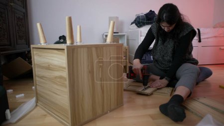 Woman assembling furniture at home with drilling machine, person moving to new home concept - putting together nightstand