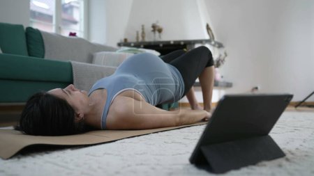 Photo for Pregnant woman practicing daily workout routine at home in front of table device at living room floor on top of Yoga mat - Royalty Free Image