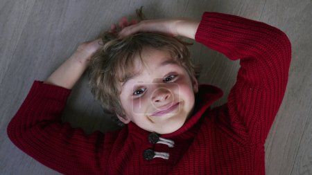 Foto de Un joven feliz sonriendo a la cámara tendido en el suelo mirando a la cámara desde abajo. Sonriente niño de 7 años con suéter rojo - Imagen libre de derechos
