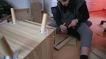 Téléchargez les photos : Femme passionnée de bricolage installant une table de chevet dans une nouvelle maison, employant une perceuse pour l'assemblage de meubles, capturant l'essence de la configuration de la maison et de l'initiative personnelle - en image libre de droit
