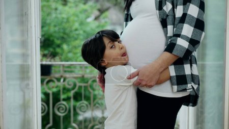Kleiner Junge zeigt Zuneigung zu ungeborenen Geschwistern, umarmt den Bauch der Mutter im warmen Zuhause auf dem Balkon