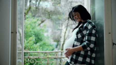 Foto de Mujer embarazada alegre cariñosamente acariciando el vientre, de pie en la ventana del balcón del apartamento con vistas panorámicas - Esencia del amor maternal, esperando al recién nacido - Imagen libre de derechos