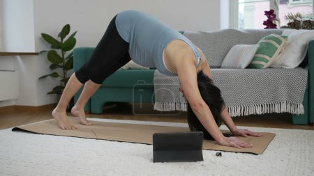 Foto de Madre embarazada comprometida en la rutina segura de acondicionamiento físico en el hogar, realizando ejercicios de levantamiento de piernas en la estera de yoga en la sala de estar, utilizando tableta digital para guiar el ejercicio - Imagen libre de derechos