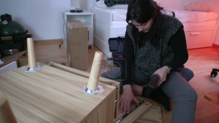 Femme passionnée de bricolage installant une table de chevet dans une nouvelle maison, employant une perceuse pour l'assemblage de meubles, capturant l'essence de la configuration de la maison et de l'initiative personnelle