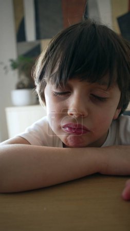 Téléchargez les photos : Un petit garçon bouleversé appuyé sur la table avec une expression de tromperie, ennuyé triste enfant de 5 ans, vidéo verticale - en image libre de droit