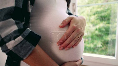 Foto de Mujer embarazada a los 8 meses, acariciando el vientre por la ventana, hogar pacífico con árboles, escena de primer plano de amor maternal y afecto - Imagen libre de derechos