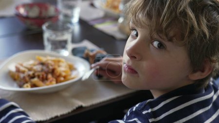 Foto de Niño girando la cabeza hacia la cámara sentado en la mesa del almuerzo sonriendo. Niño comiendo comida de pasta, vuelve a la cabeza al plato. Retrato guapo - Imagen libre de derechos