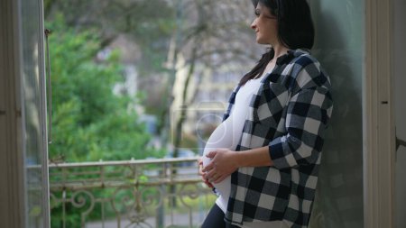 Foto de Mujer embarazada feliz acaricia suavemente el vientre de pie junto a la ventana en el balcón del apartamento con vistas - concepto de amor maternal, anticipando al bebé recién nacido - Imagen libre de derechos