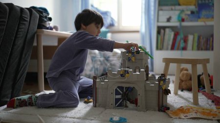 Téléchargez les photos : Petit garçon jouant dans la chambre avec château jouet à la maison portant un pyjama. Enfant absorbé dans le jeu imaginatif dans le jeu du matin - en image libre de droit