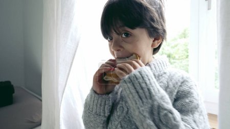 Téléchargez les photos : Happy Boy profitant d'un sandwich Close-up, petit enfant mangeant une collation riche en glucides en après-midi, debout par la fenêtre rétro-éclairée - en image libre de droit