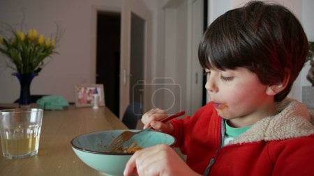 Foto de Niño disfrutando del plato de espagueti para la cena por la noche, niño caucásico de 5 años sentado a la mesa haciendo girar la pasta con tenedor, comiendo comida italiana en casa - Imagen libre de derechos