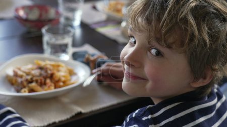 Foto de Niño girando la cabeza hacia la cámara sentado en la mesa del almuerzo sonriendo. Niño comiendo comida de pasta, vuelve a la cabeza al plato. Retrato guapo - Imagen libre de derechos