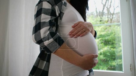 Foto de Madre embarazada a los 8 meses, acariciando tiernamente el vientre por la ventana, vista serena al hogar - Imagen libre de derechos