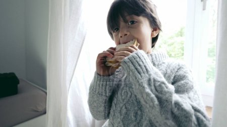 Happy Boy disfrutando de un bocadillo de cerca, un niño pequeño comiendo un bocadillo rico en carbohidratos por la tarde, de pie junto a la ventana retroiluminada