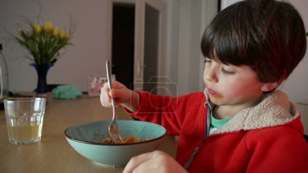 Téléchargez les photos : Enfant dégustant une assiette de spaghettis pour le souper la nuit, enfant caucasien de 5 ans assis à la table filant des pâtes à la fourchette, mangeant de la nourriture italienne à la maison - en image libre de droit