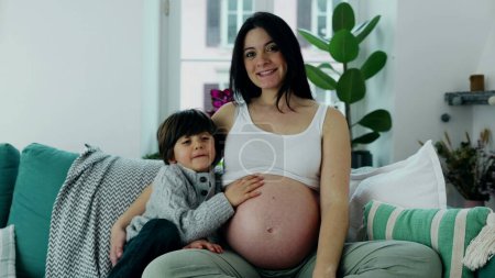Foto de Escena familiar alegre con el joven y la madre embarazada en el sofá, sonriendo a la cámara, 5 años frotando suavemente el vientre de mamá, capturando la esencia del amor maternal - Imagen libre de derechos