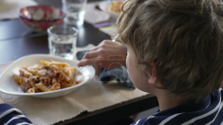 Téléchargez les photos : Jeune garçon tournant la tête vers la caméra assis à la table du déjeuner souriant. Enfant manger des pâtes alimentaires, tourne la tête vers l'assiette. Beau portrait - en image libre de droit