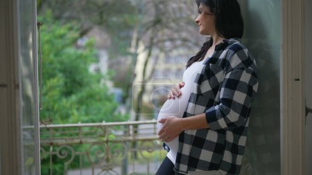 Foto de Mujer embarazada feliz acaricia suavemente el vientre de pie junto a la ventana en el balcón del apartamento con vistas - concepto de amor maternal, anticipando al bebé recién nacido - Imagen libre de derechos