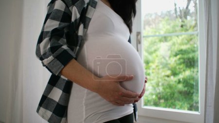 Téléchargez les photos : Femme enceinte à 8 mois, Ventre tendrement caressant par la fenêtre, Vue Sereine sur la Maison - en image libre de droit