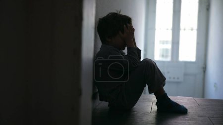 Foto de Niño pequeño que experimenta una crisis familiar, sentado en solitario en un pasillo con poca luz, cubriendo la cara con las manos para expresar profundo dolor y dolor - Imagen libre de derechos