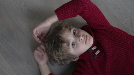 Photo for Pensive young boy plays with hair while looking at camera laid on floor looking from bellow. Child wearing red sweater with thoughtful expression, kid daydreaming - Royalty Free Image
