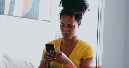 Photo for Young African American woman texting message at home seated at home sofa. One candid black latina adult girl in 20s using modern technology to communicate with friends online - Royalty Free Image