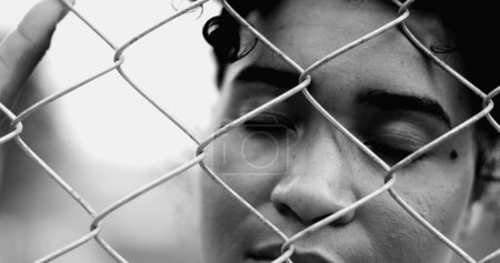 Téléchargez les photos : Une jeune femme noire enfermée penchée sur une clôture métallique fermant et ouvrant les yeux tandis que la main tient la barrière fermement luttant en silence dans un spectaculaire monochrome, noir et blanc - en image libre de droit