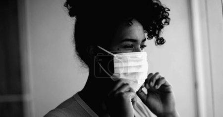 Foto de Una joven mujer negra poniendo máscara facial covid atascado en casa en intenso retrato monocromático retrato cara de cerca mirando a la ventana desde el apartamento - Imagen libre de derechos