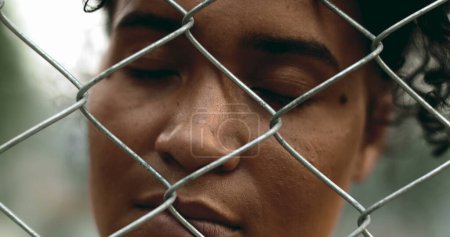 One young black woman trapped behind a fence, close-up hand and face closing eyes in solitude. 20s person struggling with mental illness behind metal barrier depicting depression