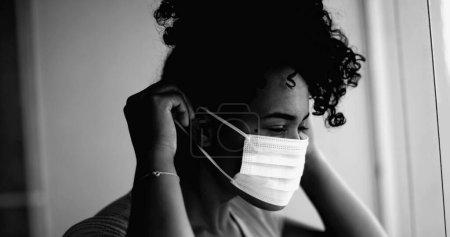 Photo for One young black woman putting covid face mask stuck at home in intense dramatic monochromatic portrait close-up face gazing at window from apartment - Royalty Free Image