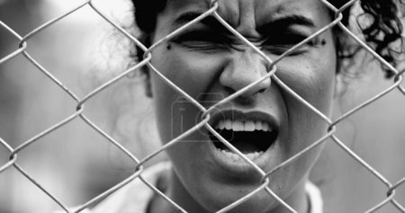 Desperate Young African American woman feeling outrage screaming with rage behind metal fence barrier frowning and open mouth screaming in anger