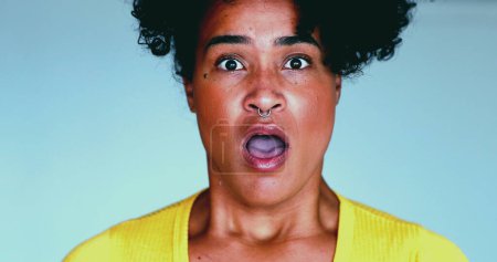 Photo for One young black woman reacts negatively to notification news looking directly at camera with open mouth and shock. Unbelief emotion of 20s person - Royalty Free Image