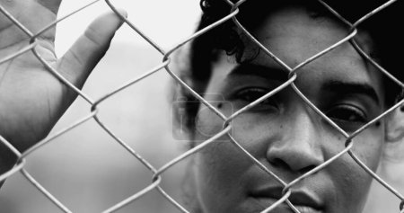 Una joven negra confinada apoyada en una valla metálica cerrando y abriendo los ojos mientras la mano sostiene la barrera firmemente luchando en silencio en dramático monocromo, blanco y negro